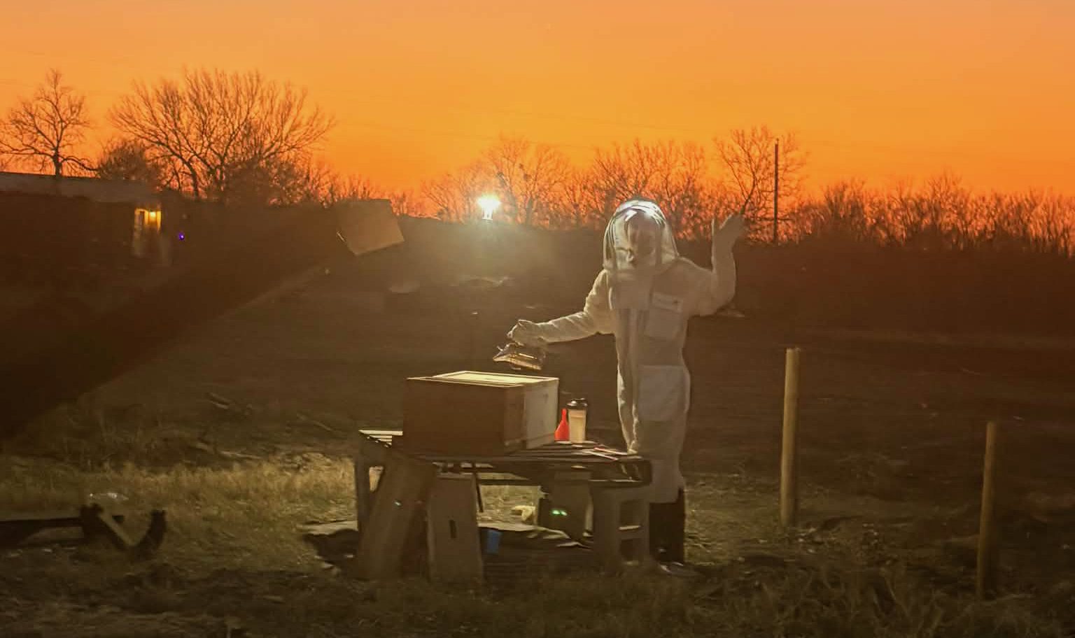 Cassie in a beekeeping suit at sunset, waving beside a hive with a smoker.